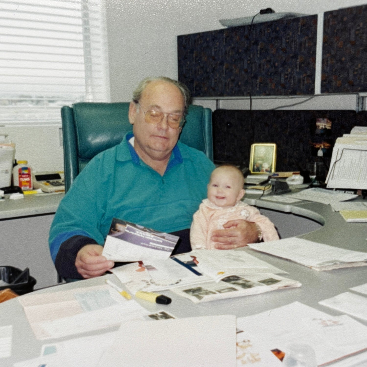 Man sitting at a desk with a baby on his lap, surrounded by papers and office supplies.