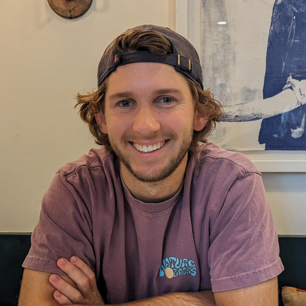 Man wearing a purple t-shirt and cap sitting at a table with a cup, smiling.