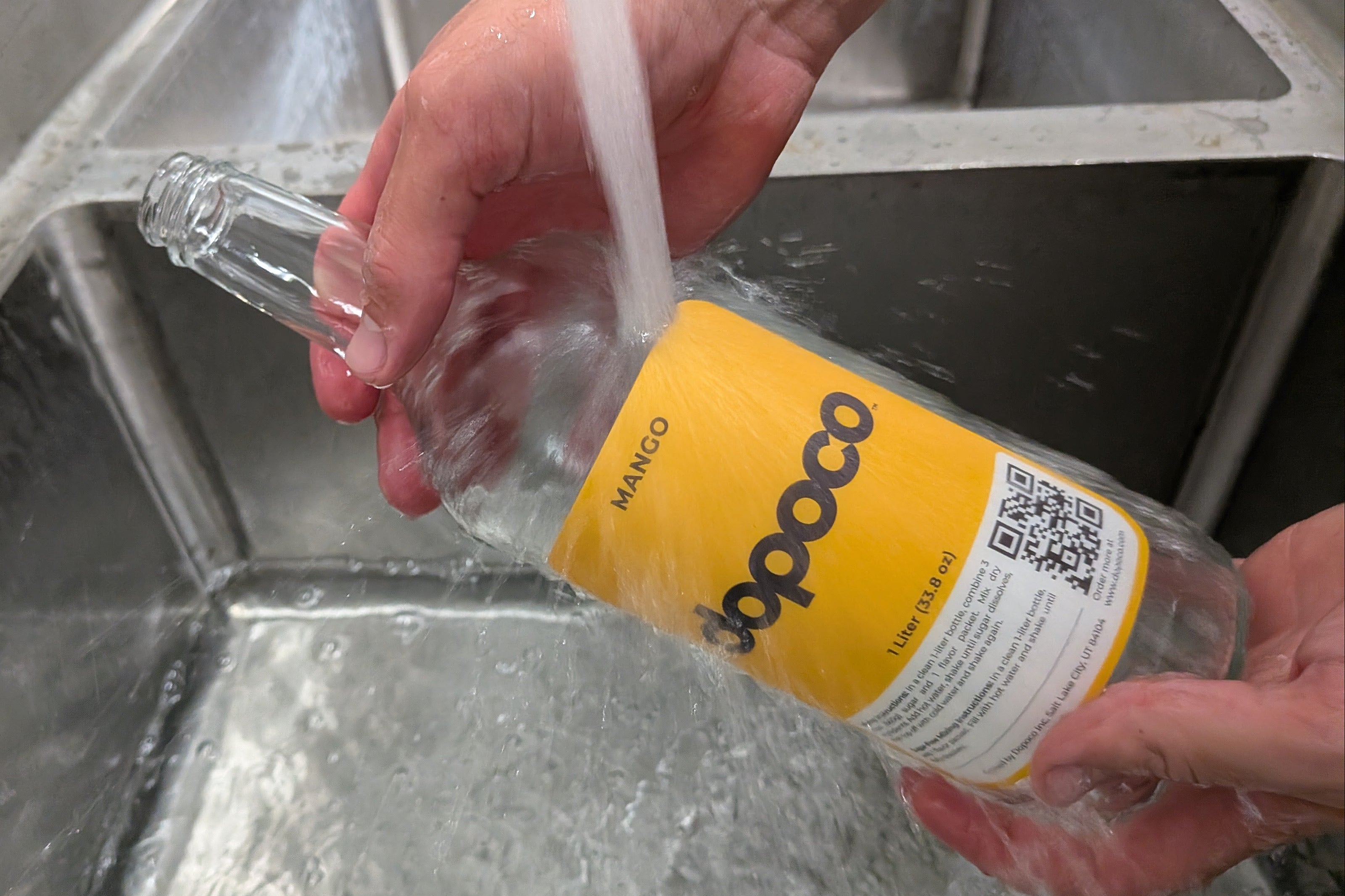 Person washing a bottle labeled 'dopoco' under running water in a sink.